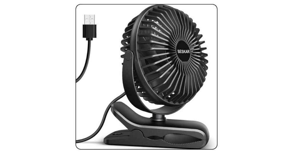 Small black USB clip-on fan clamped onto edge of college dorm desk with laptop and textbooks nearby and cord running to power strip
