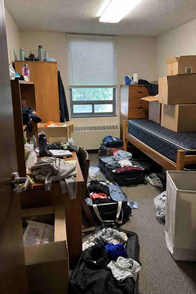 College dorm room on move-in day with open suitcases scattered across the floor clothes piled on the desk bare mattress visible and cardboard boxes stacked before any organization has started